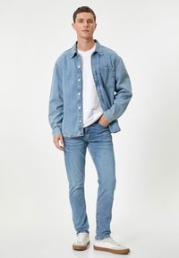 Chaqueta de mezclilla azul claro sobre una camiseta blanca, combinada con jeans azul claro y zapatillas blancas con suelas marrones. Diseño y ajuste simple.