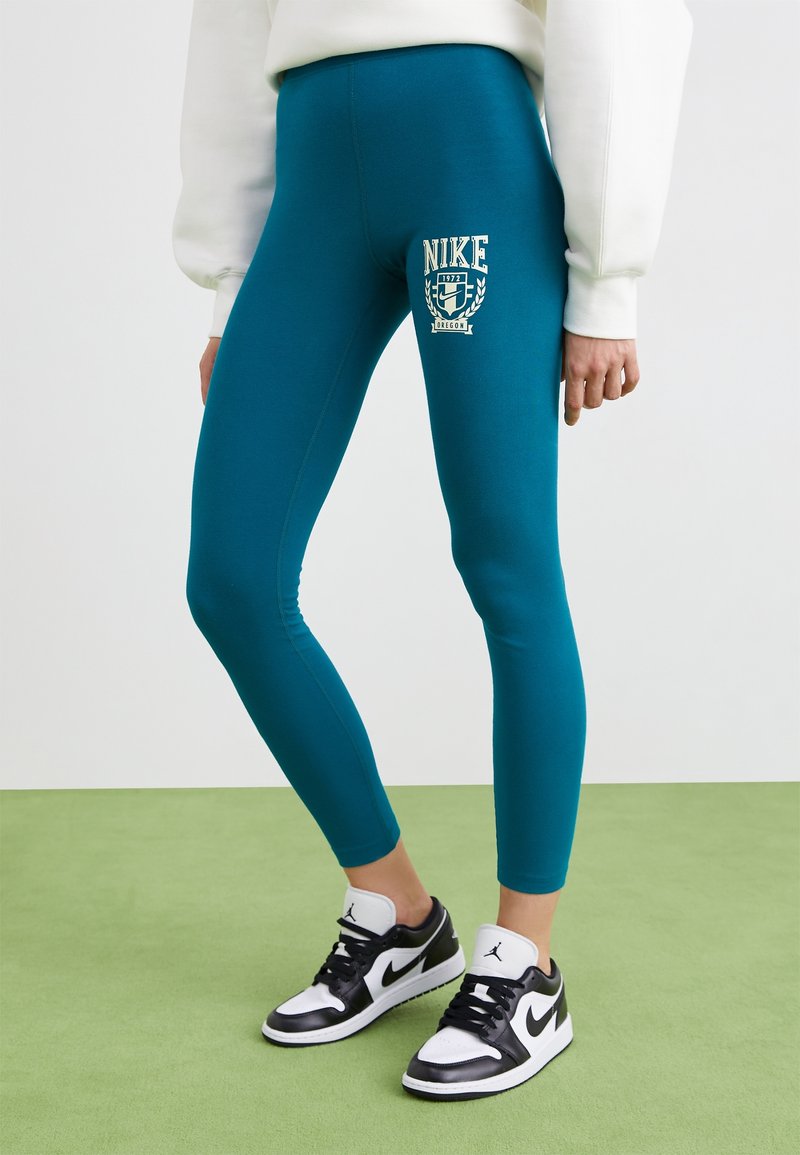 Turkosa leggings med en åtsittande design, med en vit Nike-logotyp och emblem på låret, kombinerat med svartvita sneakers.