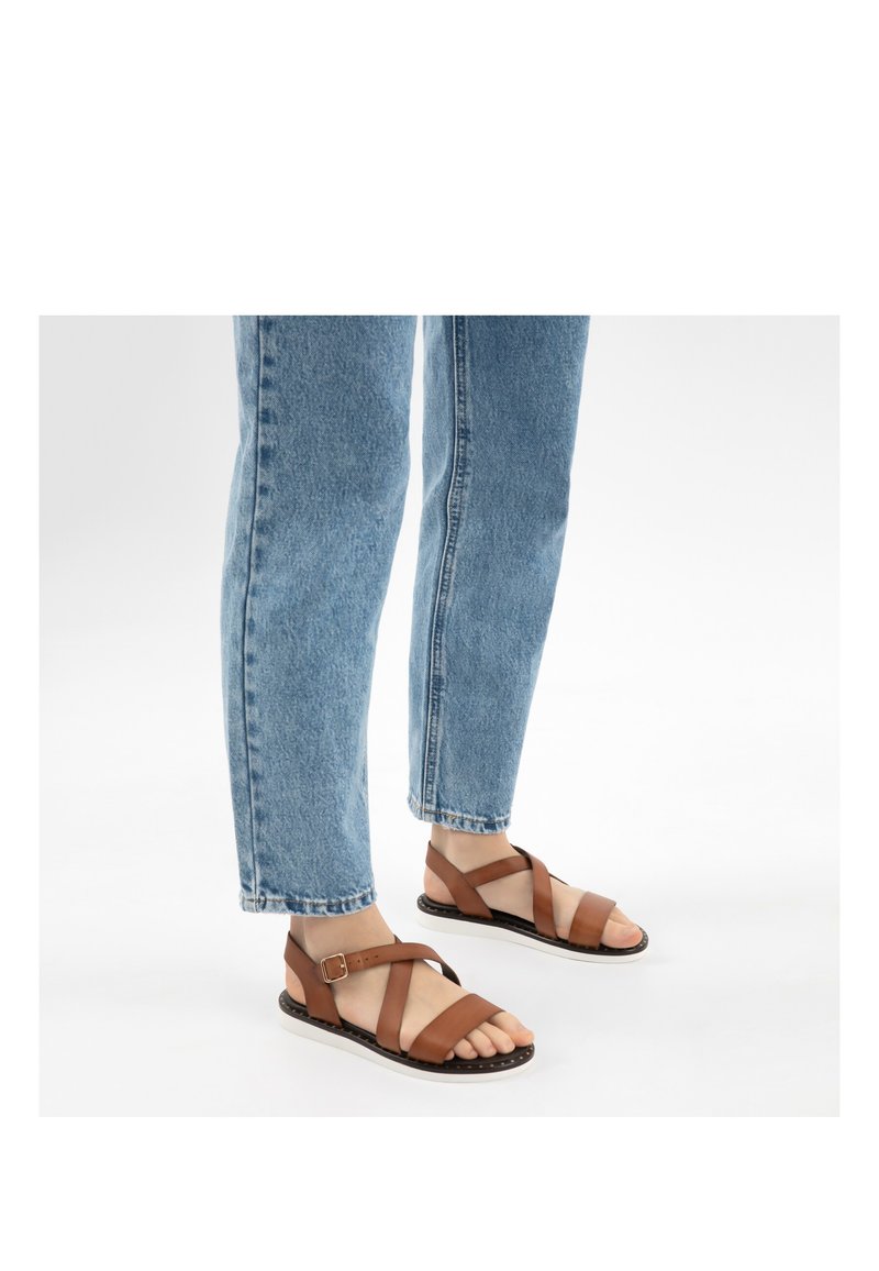 Füße, die braune Ledersandalen mit Schnallenriemen tragen, kombiniert mit blauen Jeans auf weißem Hintergrund.