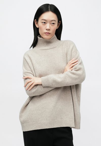 Νεαρή γυναίκα με ίσια μαύρα μαλλιά, που φοράει μπεζ πουλόβερ με ζιβάγκο και μαύρο παντελόνι, με σταυρωμένα χέρια, κοιτάζει προς τα πλάγια.