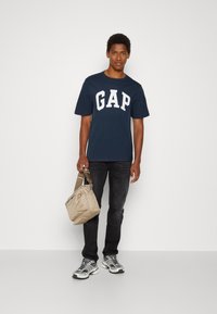 Navy T-shirt with white "GAP" logo, paired with dark jeans and grey sneakers, holding a tan bag with a short strap.