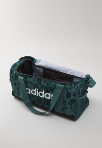 Grønn leopardmønstrete treningsbag med svarte detaljer, som har et glidelåslukket hovedrom, en side-nettlomme og en fremtredende hvit logo.