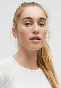 Portret osobe s dugom plavom kosom u bijelom topu, s okruglim izrezom i prozirnim akcentima na ramenima. Minimalistička estetika.