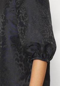 Černý top s balónovými rukávy, vyrobený z texturované tkaniny se subtilním vzorem, s jemným skladem a detaily na nařasených manžetách.