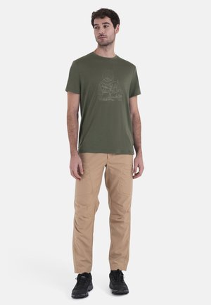 Olivgrünes T-Shirt mit einem weißen Berggrafik, kombiniert mit beigen Cargo-Hosen und schwarzen Sportschuhen. Glatte Stoffqualität mit lässigem Schnitt.