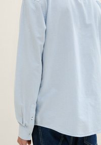 Camisa de manga larga color azul claro con finas rayas blancas, puños abotonados, corte entallado y dobladillo curvado, combinada con jeans de mezclilla oscura.