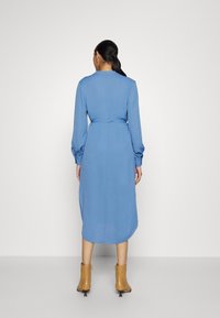 Donna con capelli scuri in un vestito midi blu a maniche lunghe e stivali alla caviglia beige, in piedi di fronte a una parete grigia chiara e semplice.
