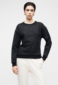 Sweatshirt preta com um padrão texturizado de formas entrelaçadas em “A”. Apresenta gola redonda e punhos canelados, combinada com calças de cor creme.