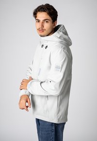 Hellgraue, wasserdichte Jacke mit Kapuze, verstellbaren Zugbändern, frontaler Reißverschluss und elastischen Bündchen. Mit einem Urban Edition-Label am Ärmel.