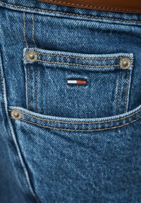 Κοντινή λήψη τσέπης από μπλε τζιν με πορτοκαλί ραφές, μεταλλικά νιβερώματα και ένα μικρό ορθογώνιο κόκκινο, λευκό και μπλε λογότυπο στο κέντρο της τσέπης.