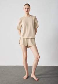 Beige bomull lediga kläder som inkluderar en lös shortärmad topp och matchande shorts med avslappnad fåll och sidoklipp. Barfota modell.