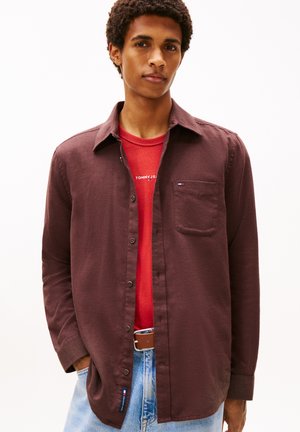 Junger Mann trägt ein offenes dunkelbraunes Hemd über einem roten Tommy Jeans T-Shirt und hellblaue Jeans mit braunem Gürtel, steht vor weißem Hintergrund.