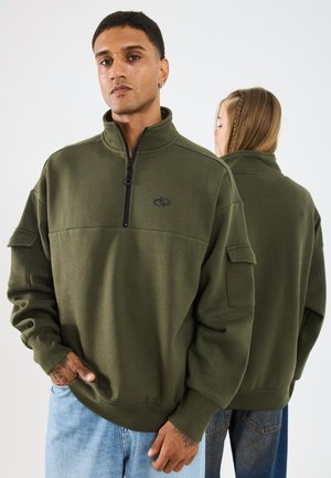 Olivroheline fleece pullover kõrge kraega, pool-zip sulgemine, eesmine tasku ja lõdva lõikega; rinna peal on logo ja soonikkoes mansetid.