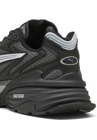 Czarne sportowe buty z siateczkową tkaniną, smukłym designem, srebrnymi akcentami oraz grubą, miękko wyściełaną podeszwą z teksturowanymi wzorami i wyraźnym logo.