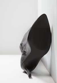 Chaussure à talon haut en cuir noir avec un bout pointu, présentant un talon fin et une semelle en caoutchouc texturé, inclinée contre une surface blanche.