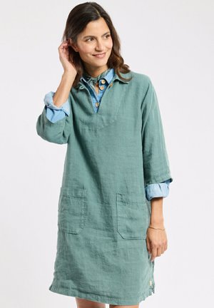 Femme portant une robe en lin à manches longues couleur sarcelle par-dessus une chemise bleue retroussée, souriante et se touchant les cheveux devant un fond clair uni.