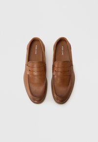 Mocasines de cuero marrón con una textura suave y detalles de costura en la parte superior, presentando una punta redondeada y un sutil logotipo en el interior.