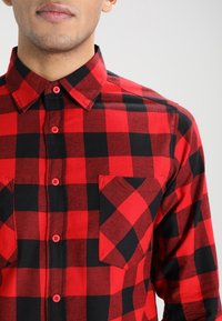 Man in een rood en zwart geruit overhemd met een kraag en een borstzak, weergegeven van de schouders tot het midden van de romp.