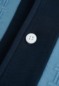 Mörkblått stickat tyg med en ljusblå accent. Har en vit knapp med "Ted Baker" präglad, placerad nära knäppningen.