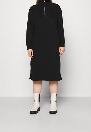 Zwarte sweatshirt jurk met een halve rits, lange mouwen en een rechte snit. Gecombineerd met lichtgekleurde enkelboots met zwarte accenten.