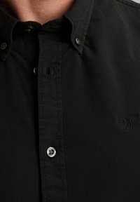 Schwarze Button-Up-Bluse mit spitzem Kragen, subtiler Textur und gesticktem Adler-Logo auf der linken Seite. Verfügt über einen Metallknopf am Kragen.