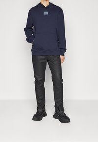 Persona con una sudadera Hugo azul marino, pantalones negros ajustados y zapatillas de senderismo negras, de pie contra un fondo blanco sencillo.