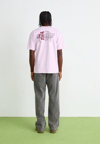 Light pink cotton t-shirt with a graphic print on the back. Paired with grey wide-leg pants and brown footwear. Simple design and relaxed fit.