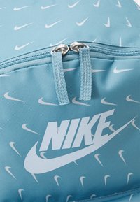 Ljusblå Nike ryggsäck med vita swoosh-mönster, med en zippered topp och dubbel dragkedjeöppning, stor vit Nike-logotyp på framsidan.