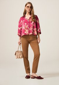 Roze bloemenblouse, bruine slim-fit broek en bordeaux rode instappers. Houdt een gestructureerde beige tas met kettinghengsel vast. Glad materiaal en platte hakken.