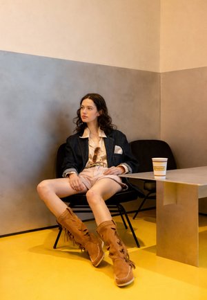 Mujer con cabello rizado sentada en una silla negra, vistiendo una chaqueta oscura, camisa beige, pantalones cortos y botas marrones con flecos en una habitación minimalista.