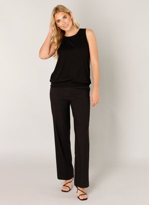 Frau mit langen blonden Haaren, die ein ärmelloses schwarzes Oberteil, schwarze, weit geschnittene Hosen und schwarze Riemchenabsätze trägt, steht vor einem schlichten hellen Hintergrund.