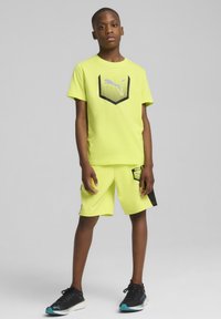 Set de tineret PUMA: cămașă scurtă, galben aprins, cu mânecă scurtă și logo negru, pantaloni scurți asortați cu accente negre și adidași negri cu detalii turcoaz.