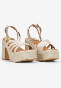 Sandalias de plataforma beige con correas entrelazadas y hebilla en el tobillo. Fabricadas con un material de cuerda texturizada, presentan un tacón grueso y una suela plana.