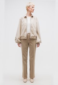 Persoană cu păr scurt blond, purtând o jachetă bej cu fermoar, top alb, pantaloni bej și pantofi bej, stând în fața unui fundal alb simplu.