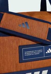 Bolsa de tela en navy y naranja con un logo de tres rayas, textura tejida y una etiqueta que presenta el texto de la marca en tres idiomas.