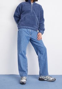 Mörkblå fleece-tröja med halv dragkedja, i kombination med ljusblå denimjeans och grå träningsskor med svarta detaljer.