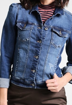 Kvinde iført en blå denimjakke over en rød- og sortstribet rullekrave, med den ene hånd holdende jakken nær taljen.
