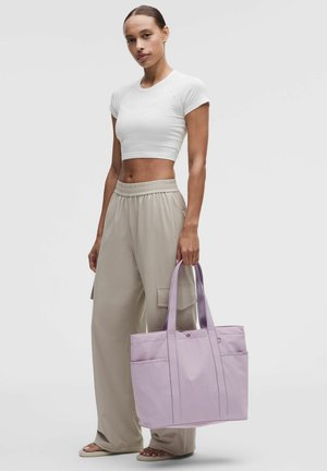 Vrouw draagt een wit cropped T-shirt en beige wijde broek, ze houdt een grote lavendelkleurige tote tas met zakken vast.
