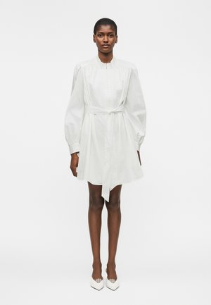 Γυναίκα που φοράει λευκό μίνι φόρεμα με μακριά μανίκια, ζώνη, φουσκωτά μανίκια και μυτερές λευκές γόβες, στέκεται μπροστά σε απλό φόντο.