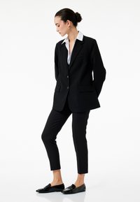 Sort blazer med hvid krave, skræddersyet fit og forlommer, sammen med sorte slim-fit bukser og skinnende sorte loafers.