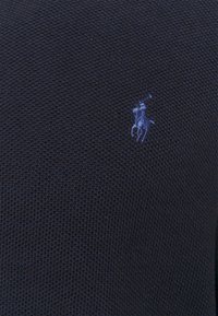 Mörkblå stickad polo tröja med ett strukturerat mönster, med en liten ljusblå broderad logotyp av en häst och ryttare på vänster bröst.