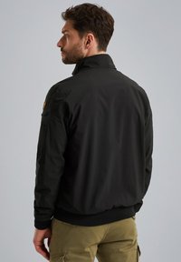 Schwarze Bomberjacke mit hohem Kragen, gerippten Bündchen und seitlichem Taschen-Detail. Glatte Textur und stromlinienförmiges Design.