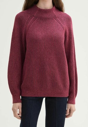Femme portant un pull à manches longues côtelé, rose foncé avec un col montant, associé à un jean bleu foncé.