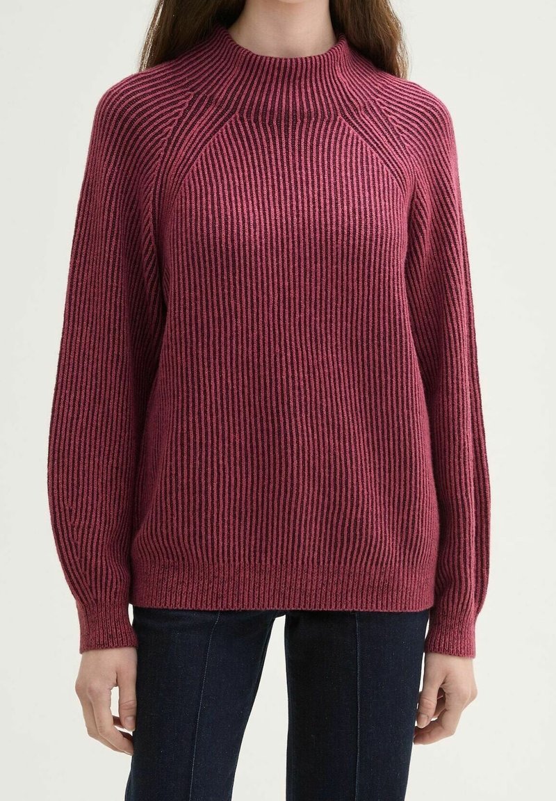Femme portant un pull à manches longues côtelé, rose foncé avec un col montant, associé à un jean bleu foncé.