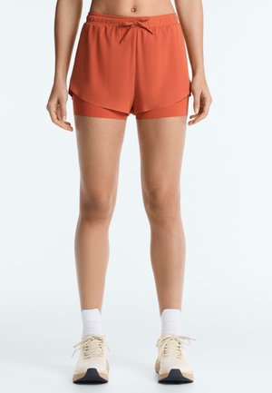 Kvinde iført orange lagdelte sportsshorts med snøre, hvide sokker og beige løbesko, stående mod en ensfarvet baggrund.