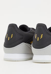 Schwarze und graue Sneakers mit einem Obermaterial aus Wildleder und Mesh, weißer Sohle und goldenen Akzenten an der Ferse sowie einem markanten Logo-Detail.