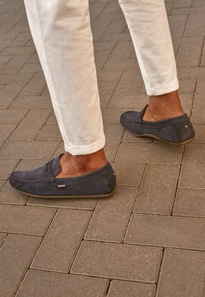 Persoon draagt witte broek en donkerblauwe suède loafers en loopt over een gestructureerde bruine bestrating.