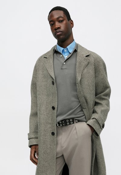 Sivi i crni kaput s uzorkom kariranog zubca, nosi se preko sive polo majice i svijetloplave košulje s ovratnikom. Hlače su bež boje s crnim remenom koji ima okrugle detalje.