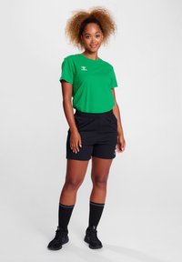 Zielona bawełniana koszulka z logo, zestawiona z czarnymi szortami na gumce w pasie. Czarne skarpetki do kolan i trampki dopełniają strój.