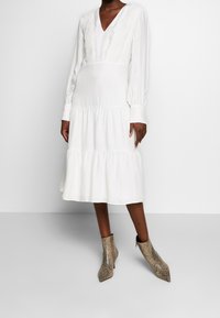 Biała sukienka o średniej długości z długimi rękawami, dekoltem w serek, teksturowaną koronkową wstawką i tiered hem. Połączona z cholewkami na wskazującym obcasie w wzór skóry węża.